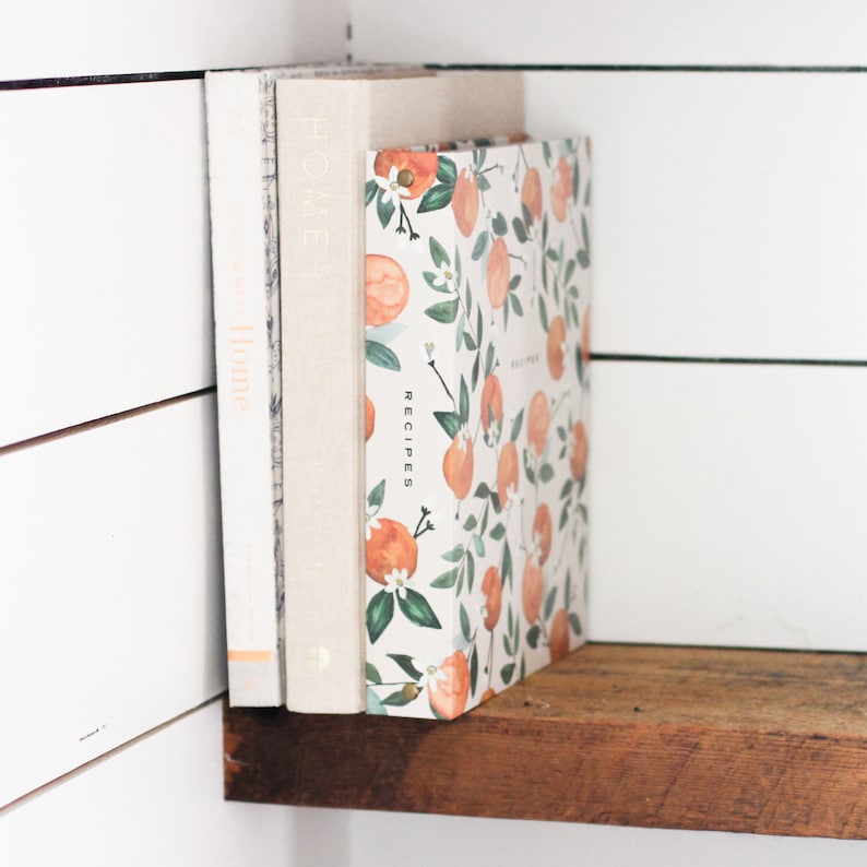 May include: A white shelf with two books stacked on top of each other. The top book has a white cover with a floral pattern of orange blossoms and oranges. The text "RECIPES" is printed vertically on the front cover. The bottom book has a white cover with the text "HOME" printed vertically on the front cover.