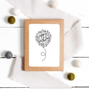 May include: A white card with a black and white illustration of a balloon with the text "happy birthday" inside. The card is on a brown background with white and green decorative balls.