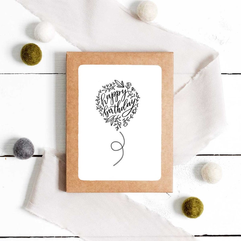 May include: A white card with a black and white illustration of a balloon with the text "happy birthday" inside. The card is on a brown background with white and green decorative balls.