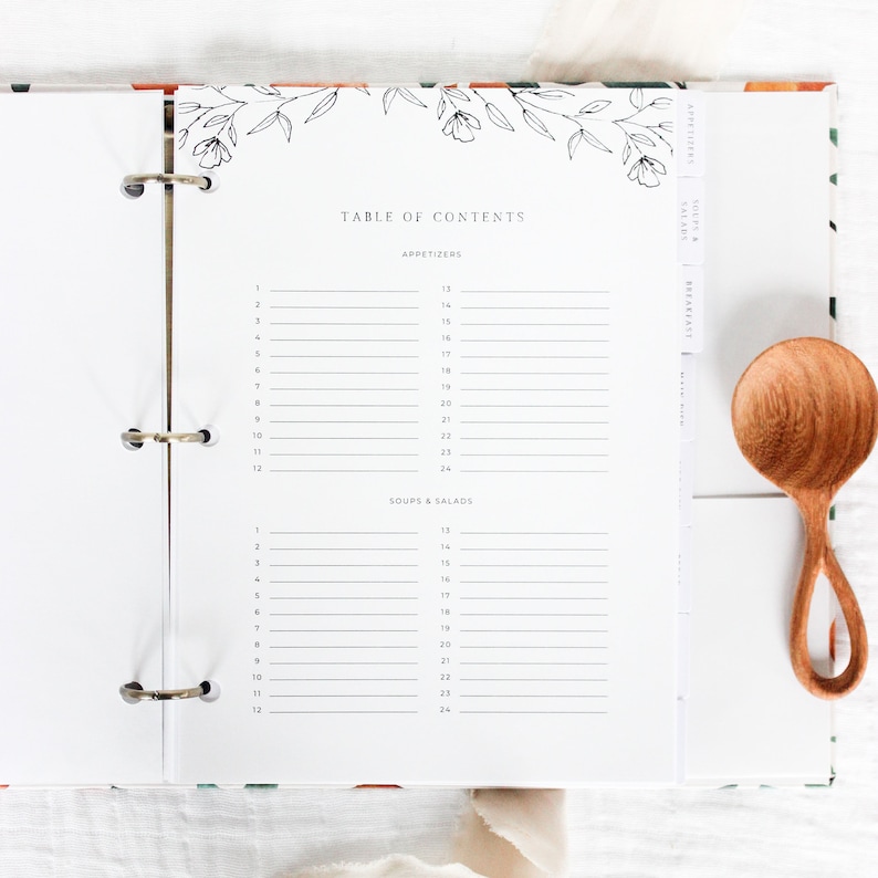 May include: A white binder with a floral design on the cover. The binder has a table of contents with sections for appetizers, soups & salads, and breakfast. The binder is open to the page with the table of contents.