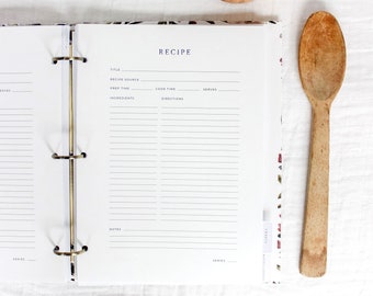 May include: Open recipe binder with gold rings, featuring a "Recipe" page with fill-in fields. A wooden spoon rests beside the open book. The binder has a floral pattern on the edges. A small, brown ceramic bowl is in the background.