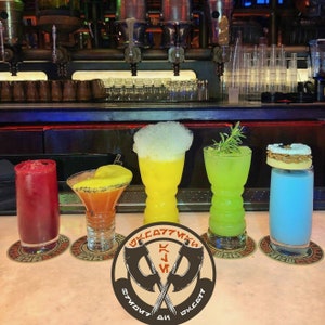 May include: Five colorful cocktails in different glasses on a bar counter with a coaster under each glass. The coasters have a design of crossed axes with the text "Oga's Cantina" in a circle around the axes.