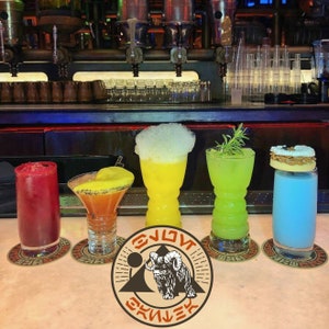 May include: Four colorful cocktails in different glasses on a bar counter. The cocktails are a red, yellow, green, and blue. The bar counter is made of a light brown material. The cocktails are arranged in a row from left to right. The background is a dark brown bar with a metal bar rail. The text "Enjoy the galaxy" is printed on a round sticker in the foreground.