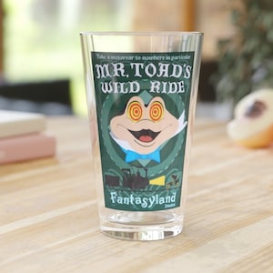 May include: A clear glass with a green, white, and blue graphic of Mr. Toad's Wild Ride from Fantasyland at Disneyland. The graphic features Mr. Toad with his eyes wide open and a big smile.