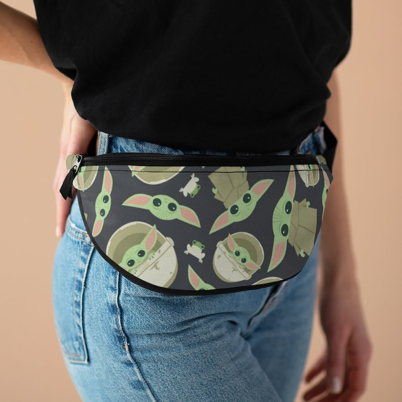 Puede incluir: Una ri&ntilde;onera negra con un patr&oacute;n repetido de personajes de beb&eacute; Yoda verdes sobre un fondo gris oscuro. La ri&ntilde;onera se lleva alrededor de la cintura de una persona que lleva pantalones vaqueros azules.