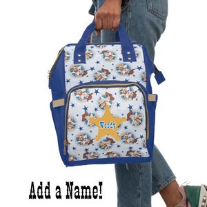 May include: A blue and white backpack featuring a cartoon character design. The backpack has a large front pocket with a star-shaped graphic and the name "Woody". The bag has blue handles and trim, and the text "Add a Name!" is at the bottom.