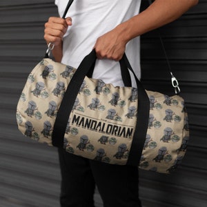 Puede incluir: Una bolsa de lona de color beige con una correa negra y un patr&oacute;n repetido de un peque&ntilde;o extraterrestre verde y un personaje mandaloriano. La bolsa tiene el texto "The Mandalorian" impreso en la parte delantera.