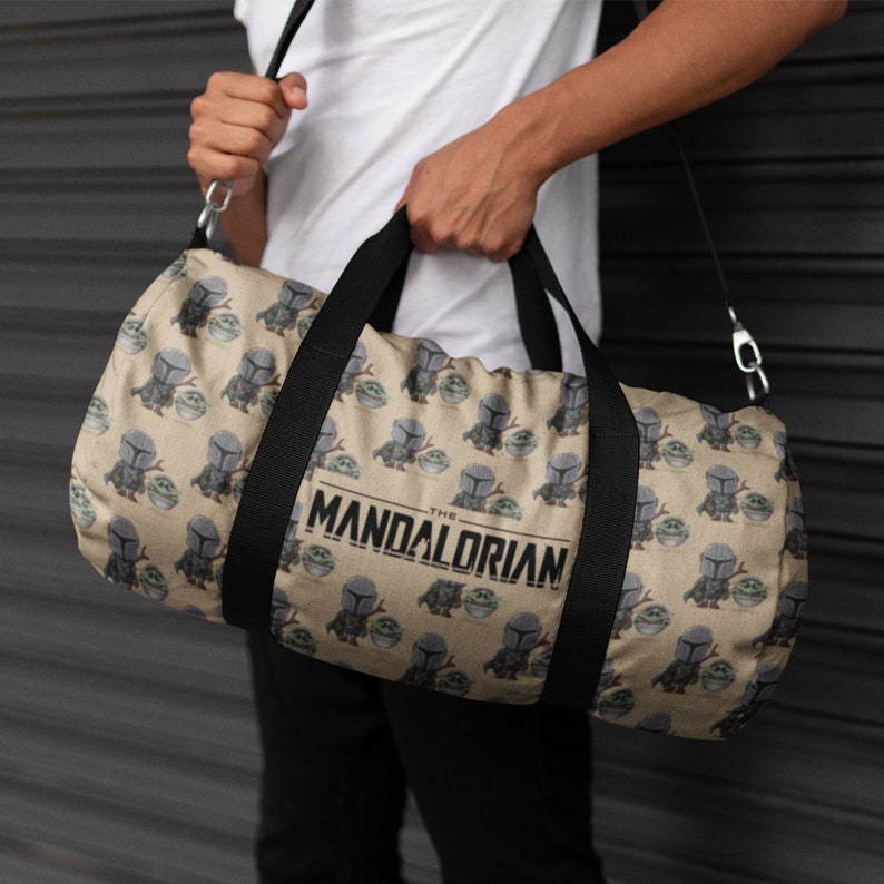 Puede incluir: Una bolsa de lona de color beige con una correa negra y un patr&oacute;n repetido de un peque&ntilde;o extraterrestre verde y un personaje mandaloriano. La bolsa tiene el texto "The Mandalorian" impreso en la parte delantera.