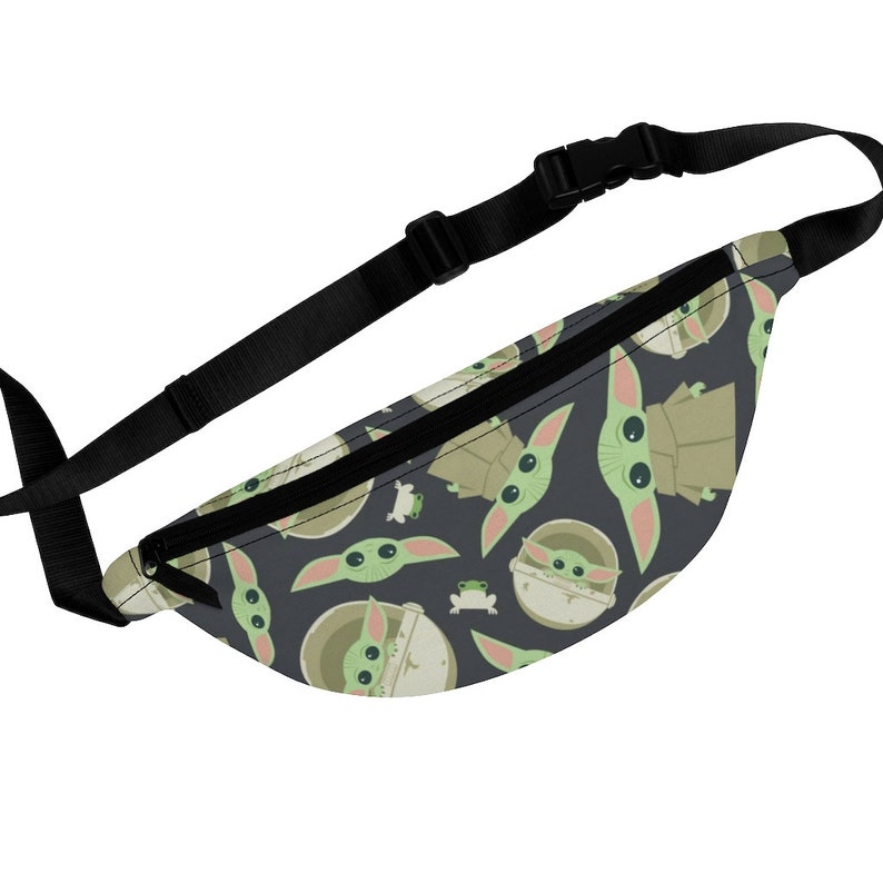 Puede incluir: Una ri&ntilde;onera negra con una correa ajustable. La ri&ntilde;onera presenta un patr&oacute;n repetido de personajes de beb&eacute; Yoda verdes sobre un fondo gris oscuro.