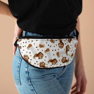 May include: A white fanny pack with a brown and white repeating pattern of a cartoon dog with a spring. The dog is wearing a brown collar and has a brown nose and eyes. The fanny pack has a black zipper and a black strap.