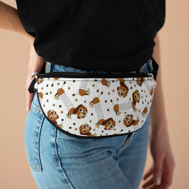 May include: A white fanny pack with a brown and white repeating pattern of a cartoon dog with a spring. The dog is wearing a brown collar and has a brown nose and eyes. The fanny pack has a black zipper and a black strap.