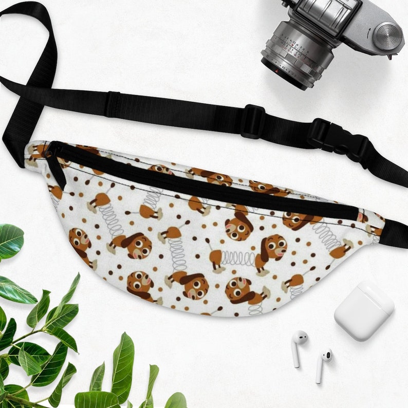 May include: A white fanny pack with a black adjustable strap. The fanny pack has a repeating pattern of brown cartoon dogs with springs on their tails and brown dots.