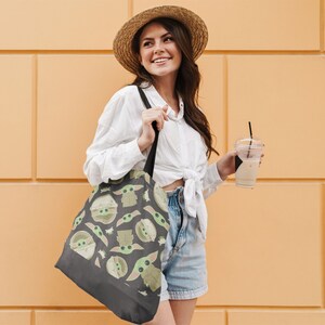 Puede incluir: Una mujer con una camisa blanca y pantalones cortos de mezclilla lleva una bolsa de mano negra con un patr&oacute;n verde y marr&oacute;n de personajes de dibujos animados. La bolsa tiene una correa negra y la mujer sostiene una taza de caf&eacute; helado.