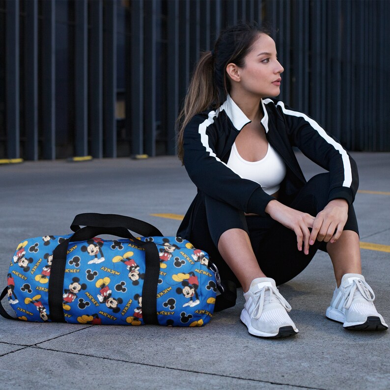 Puede incluir: Una bolsa de lona azul y amarilla con estampado de Mickey Mouse. La bolsa tiene correas negras y cierre de cremallera.