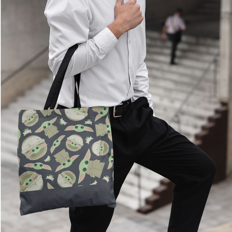 Puede incluir: Una bolsa de tela gris oscuro con un patr&oacute;n repetido de personajes de dibujos animados verdes y marrones. Los personajes tienen ojos grandes y llevan t&uacute;nicas marrones con capucha.