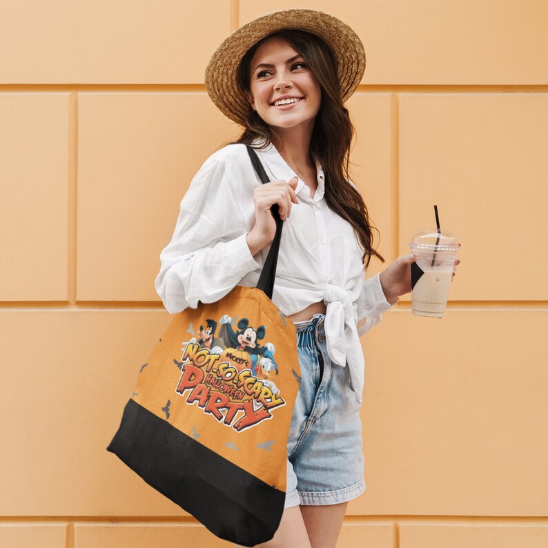 May include: A woman wearing a white shirt and denim shorts is holding a brown tote bag with a Mickey Mouse Halloween design. The bag says "Mickey's Not-So-Scary Halloween Party".