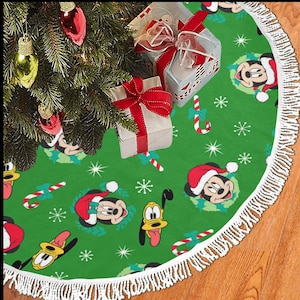 May include: A green Christmas tree skirt featuring Mickey Mouse and Pluto in Santa hats, candy canes, and snowflakes. The skirt has a white fringe border and is placed around a decorated Christmas tree with wrapped gifts.