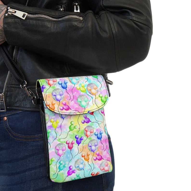 Op de afbeelding: Een witte crossbody tas met een kleurrijk aquarelpatroon van Mickey Mouse ballonnen. De tas heeft een klep sluiting en een lange verstelbare band.