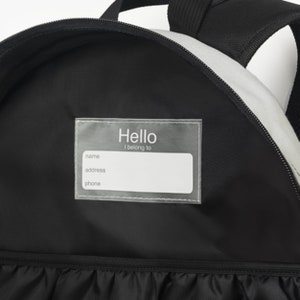 Puede incluir: Mochila negra con una etiqueta transparente de pl&aacute;stico que dice "Hello I belong to" con espacios para el nombre, la direcci&oacute;n y el n&uacute;mero de tel&eacute;fono.