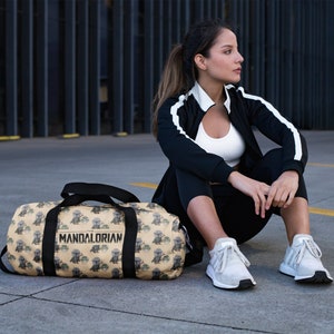 Puede incluir: Una bolsa de lona beige con una correa negra y un patr&oacute;n repetido del personaje "The Child" de la serie de televisi&oacute;n "The Mandalorian". La bolsa tiene el texto "The Mandalorian" impreso en la parte delantera.