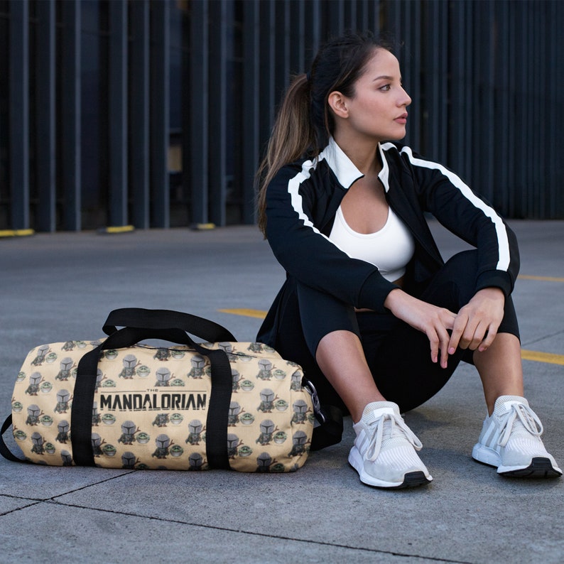 Puede incluir: Una bolsa de lona beige con una correa negra y un patr&oacute;n repetido del personaje "The Child" de la serie de televisi&oacute;n "The Mandalorian". La bolsa tiene el texto "The Mandalorian" impreso en la parte delantera.