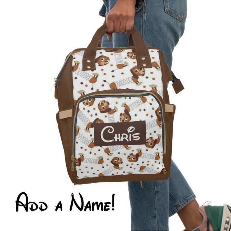 May include: Brown and white diaper bag with a repeating pattern of brown cartoon dogs with springs. The bag has a brown leather trim and a brown name tag with the name "Chris" on it.