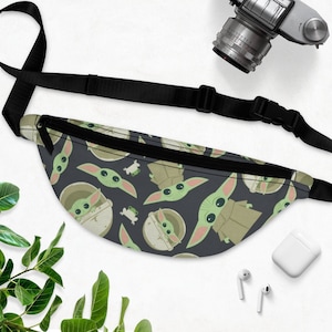 Puede incluir: Una ri&ntilde;onera negra con una correa ajustable. La ri&ntilde;onera tiene un patr&oacute;n repetido de un personaje alien&iacute;gena verde sobre un fondo azul oscuro.