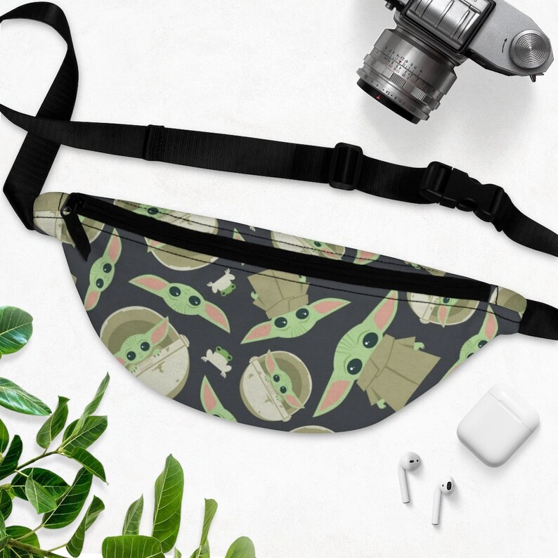 Puede incluir: Una ri&ntilde;onera negra con una correa ajustable. La ri&ntilde;onera tiene un patr&oacute;n repetido de un personaje alien&iacute;gena verde sobre un fondo azul oscuro.