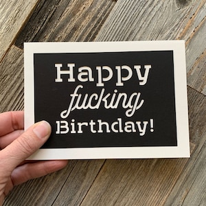 May include: A black card with white text that reads "Happy fucking Birthday!" The card is held in a hand.
