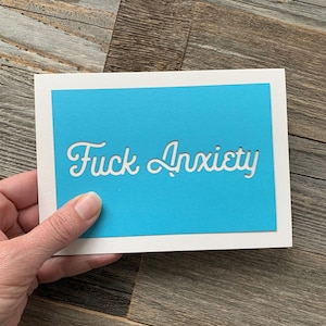 May include: A white greeting card with a blue center, featuring the words "Fuck Anxiety" cut out in a cursive font. The card is held against a wooden background.