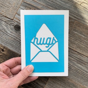 May include: A white card with a blue background and a white envelope cut out of the background. The word "hugs" is cut out of the envelope and is visible through the opening.