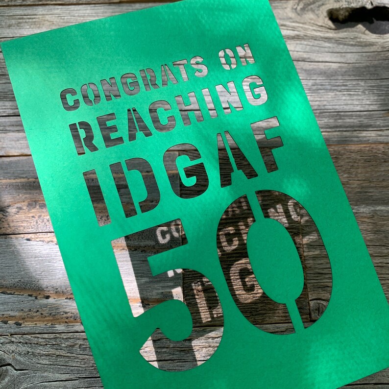 May include: A green card with cut-out text that reads "CONGRATS ON REACHING IDGAF 50". The card is angled on a wooden surface, with the text's shadows visible. The number 50 is prominently displayed.
