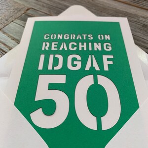 May include: A greeting card with a green background and white text that reads "CONGRATS ON REACHING IDGAF 50". The card is inside a white envelope.