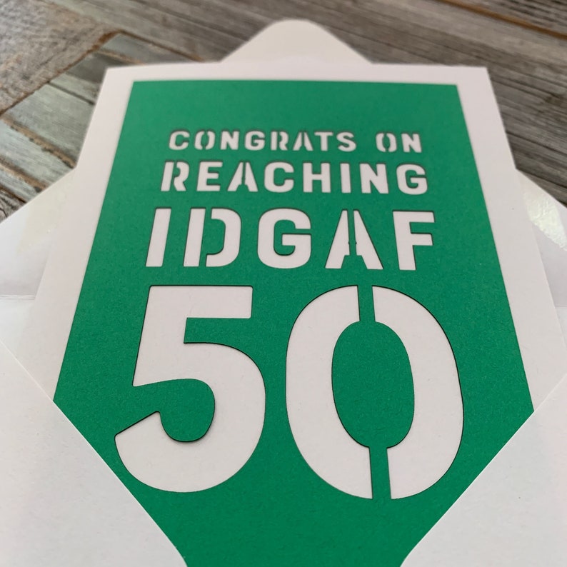 May include: A greeting card with a green background and white text that reads "CONGRATS ON REACHING IDGAF 50". The card is inside a white envelope.
