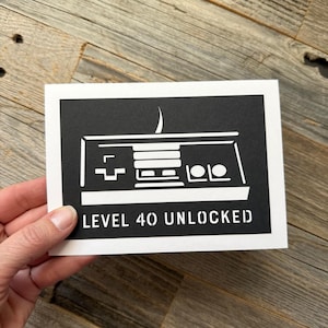 May include: A rectangular white greeting card featuring a black center panel with a white graphic illustration of a retro video game controller. Below the controller, the text reads LEVEL 40 UNLOCKED in a bold, stencil-style white font.