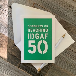May include: A greeting card with a green background and white text that reads "CONGRATS ON REACHING IDGAF 50". The card is inside a white envelope, with a pen featuring a white and gold polka dot pattern.