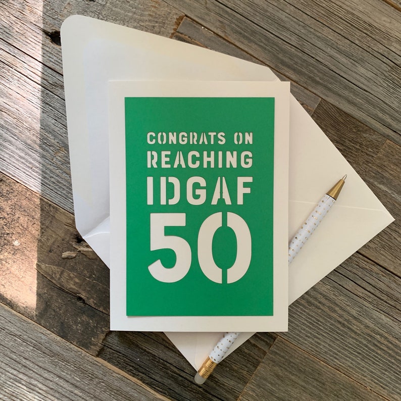 May include: A greeting card with a green background and white text that reads "CONGRATS ON REACHING IDGAF 50". The card is inside a white envelope, with a pen featuring a white and gold polka dot pattern.
