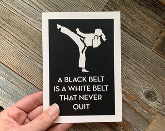 May include: A black and white motivational quote with a silhouette of a person in a karate pose. The quote reads "A black belt is a white belt that never quit."