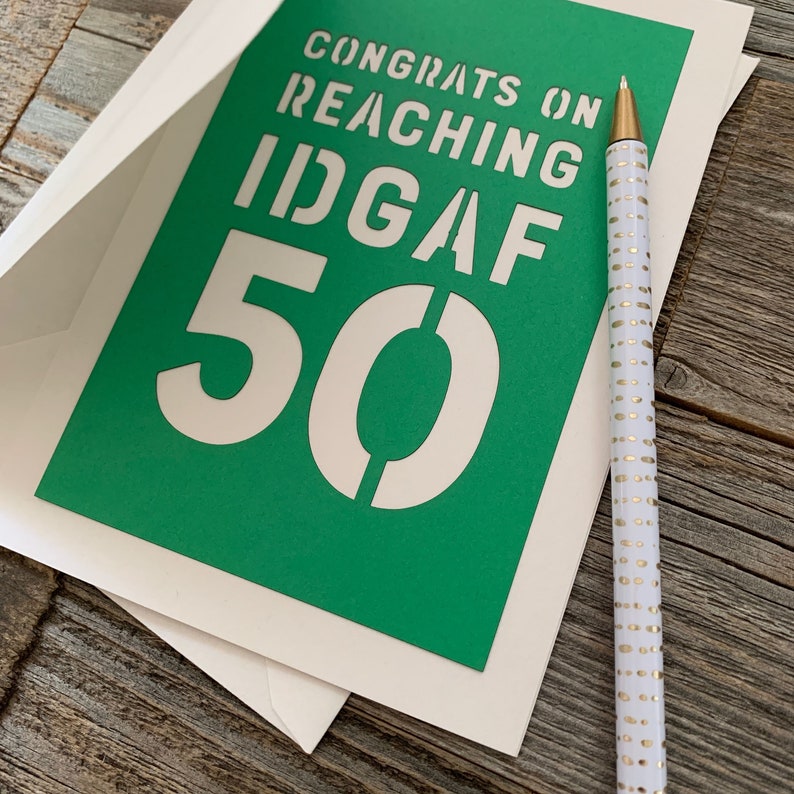 May include: A greeting card with the text "CONGRATS ON REACHING IDGAF 50" in white on a green background. The card is inside a cream-colored envelope. A white pen with gold dots rests on the card.