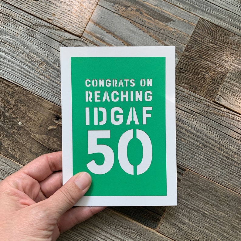May include: A white greeting card with a green background and the text "CONGRATS ON REACHING IDGAF 50" in white stencil font. The card is held by a hand against a wooden background.