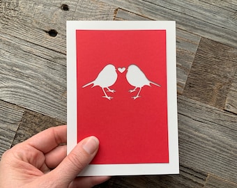 May include: A white card with a red background. Two white birds are cut out of the card, facing each other with a small heart between them.