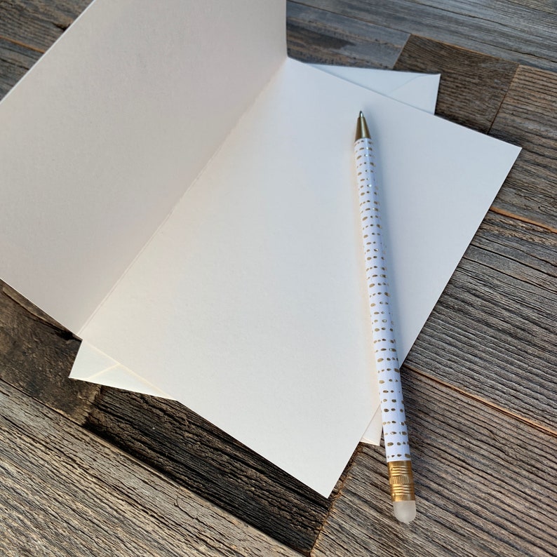 May include: An open, blank greeting card with a matching envelope and a white pencil with gold dot pattern. The pencil has a gold tip and eraser. The card and envelope are white, and the background is a weathered wood surface.