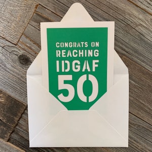 May include: A greeting card with the text "CONGRATS ON REACHING IDGAF 50" in white stencil font on a green background. The card is inside a white envelope, set against a weathered wood surface.