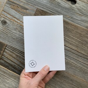 May include: A white card with rounded corners is held in a hand. The card has a small black logo and text in the lower left corner. The background is a weathered wood surface.