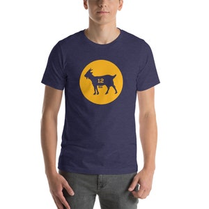 Puede incluir: Una camiseta azul marino con un círculo amarillo que presenta una silueta negra de una cabra con el número 12 en su interior. La cabra está etiquetada como "Tex" debajo del número.