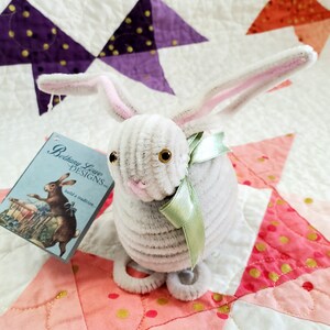 May include: A gray and white bunny rabbit made of pipe cleaners with a green ribbon around its neck. The bunny is sitting on a pink and white quilt with gold polka dots. The bunny has brown eyes and a pink nose. The bunny is holding a tag that says "Bethany Lowe Designs - build a tradition..."