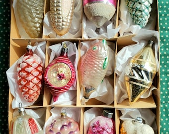 May include: A collection of twelve vintage glass Christmas ornaments arranged in a cardboard box with white tissue paper. The ornaments feature various shapes including pinecones, a fish, and spheres, with metallic, glittered, and painted finishes.