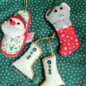 May include: A set of four hanging ornaments including a Santa figure in a holly-patterned slipper, a gray mouse in a red snowflake-patterned stocking, and two white felt ice skates with green sequins, gold trim, and small green pom-poms.