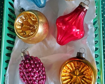 May include: A collection of vintage Christmas ornaments in a green tray. The ornaments are various shapes and colors, including blue, gold, red, and purple. Some have textured surfaces, and others have starburst or floral designs. They are arranged on white tissue paper.