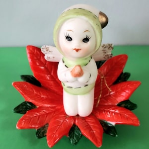 May include: A ceramic Christmas angel figurine with a white body, green hood, and red poinsettia base. The angel has a serene expression, holding its hands together in prayer. The wings are white with gold accents.